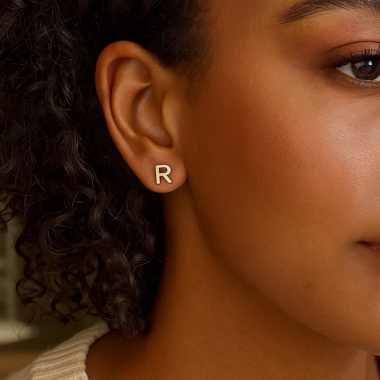 Child wearing small gold initial R stud earring, close‑up with soft lighting.