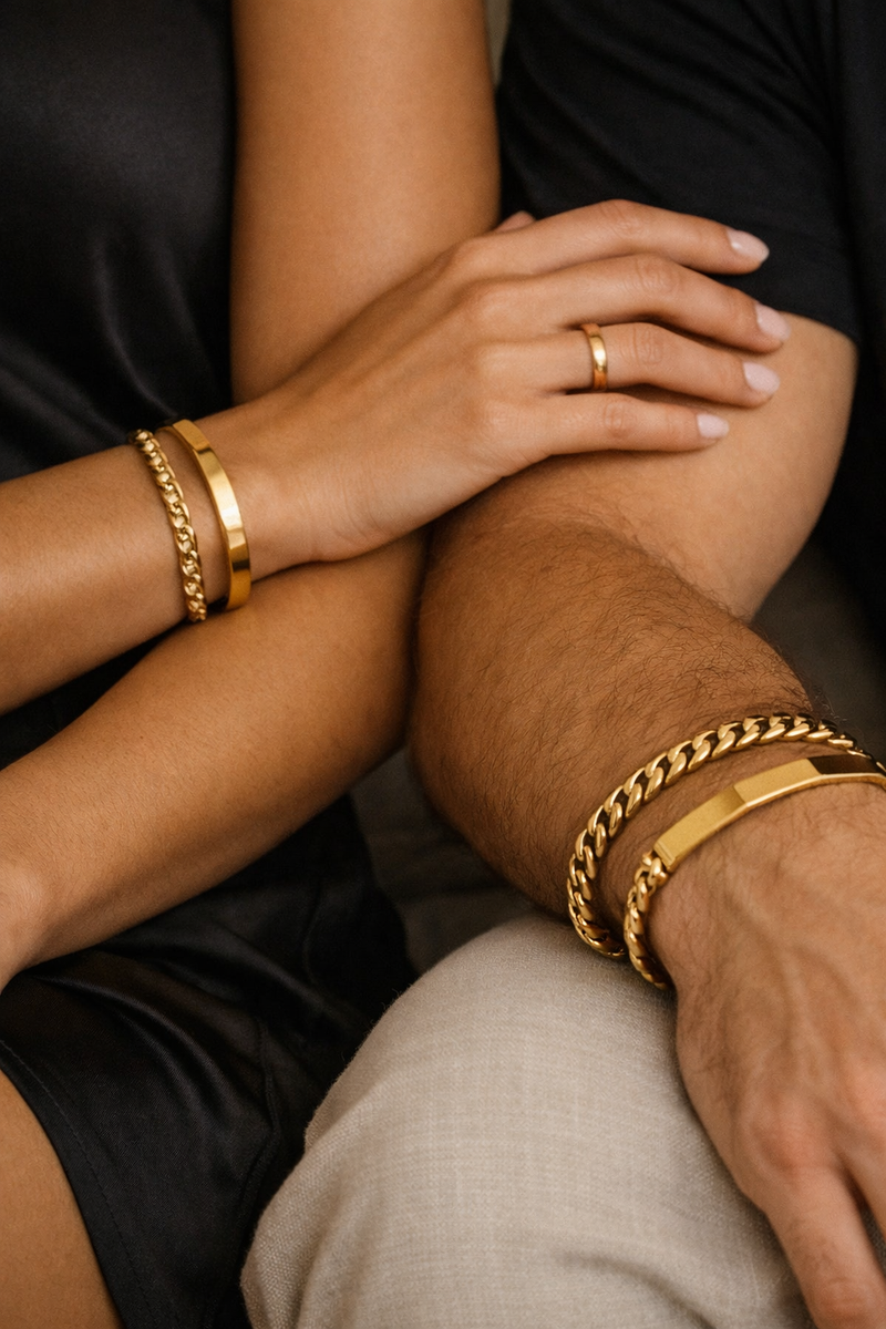 Male and female wrists wearing gold bangles and Cuban link bracelets, timeless luxury.