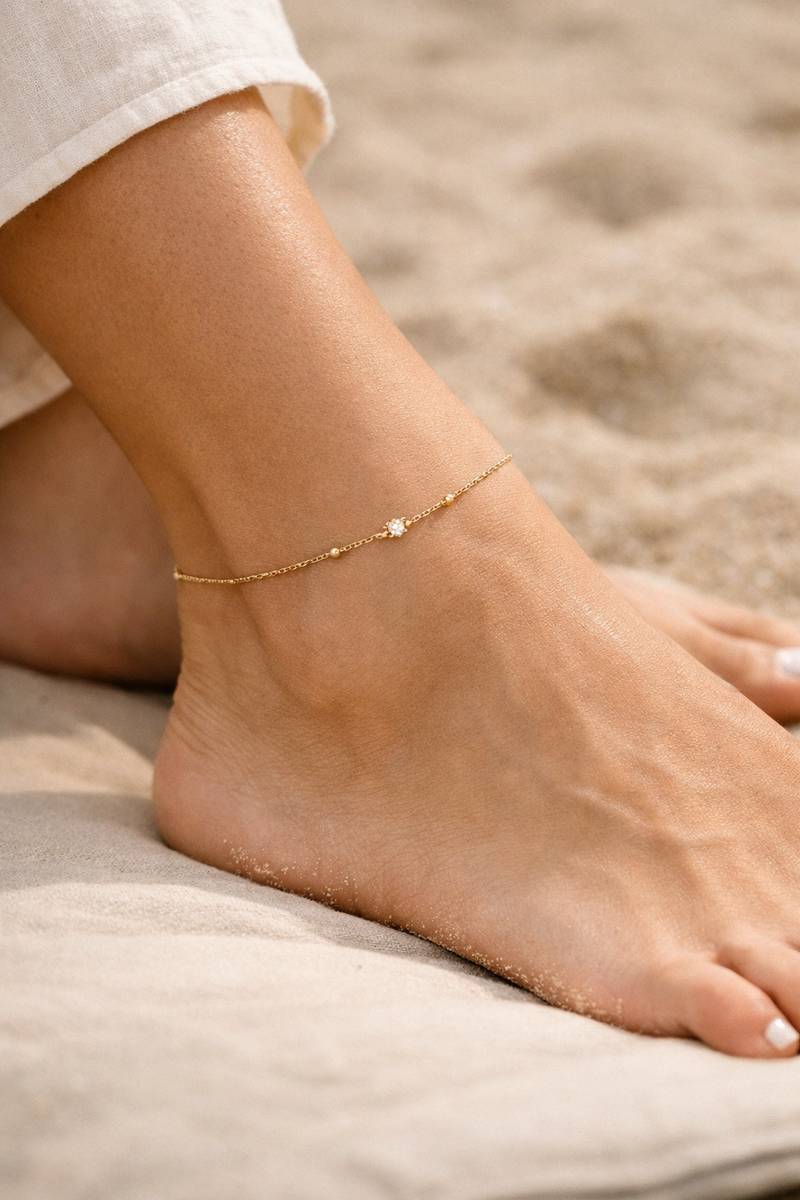 Delicate gold anklet on woman’s ankle, beach‑tone background, minimalist luxury